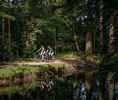 Zwei Personen fahren mit ihren Fahrrädern an einem Fluss entlang 